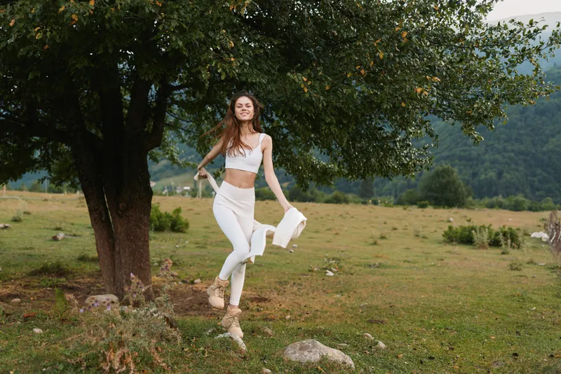 Young woman in white athleisure leaps across a sunlit field, expressing carefree energy. Natural scenery with trees and hills, lifestyle, wellness, and movement.