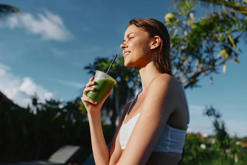 Young woman outdoors enjoying green smoothie, relaxed posture, sunny day, casual white top, natural background with trees and blue sky, healthy lifestyle concept