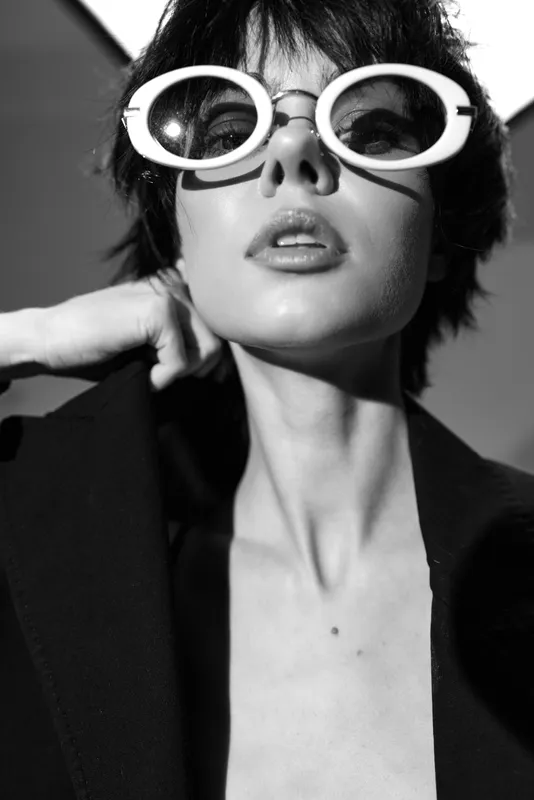 Black and white portrait of stylish woman wearing large round sunglasses and a dark jacket, expressing confidence and modern fashion.
