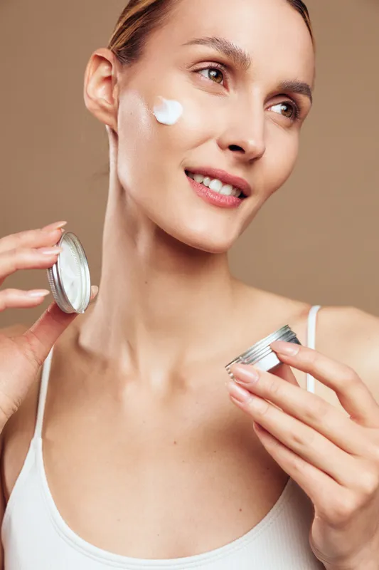 A young brunette woman applies cream from a small jar on her cheek, performing a calm skincare routine in soft studio light for a clean, modern portrait.