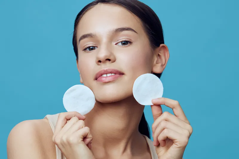 Young woman holding facial pads against a blue background, showcasing skincare routine and healthy beauty practices with a calm and confident expression