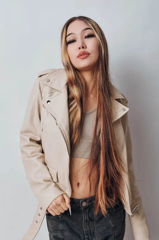 Young woman with long straight hair wearing beige leather jacket and crop top standing against light background with confident expression and casual style in studio portrait.