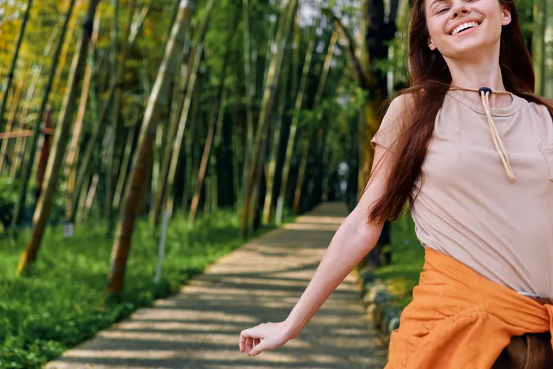 Woman smile park path walking joy nature trees, carefree young woman strolling along tree-lined walkway with jacket tied at waist, casual outfit, long hair and sunny relaxed expression.