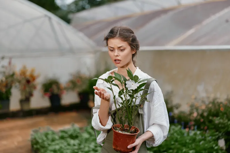 Concerned young woman in gardening attire examining a plant in a greenhouse, displaying emotions of curiosity and care for her green thumb