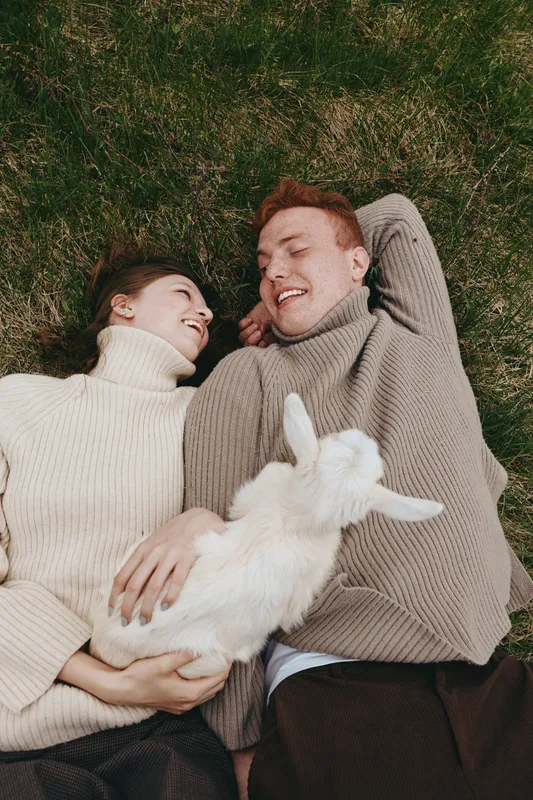 A couple lie on the grass in matching sweaters, a playful goat kid snuggled between them, sharing warm smiles in a bright outdoor moment. Concept: relaxed lifestyle and animal companionship