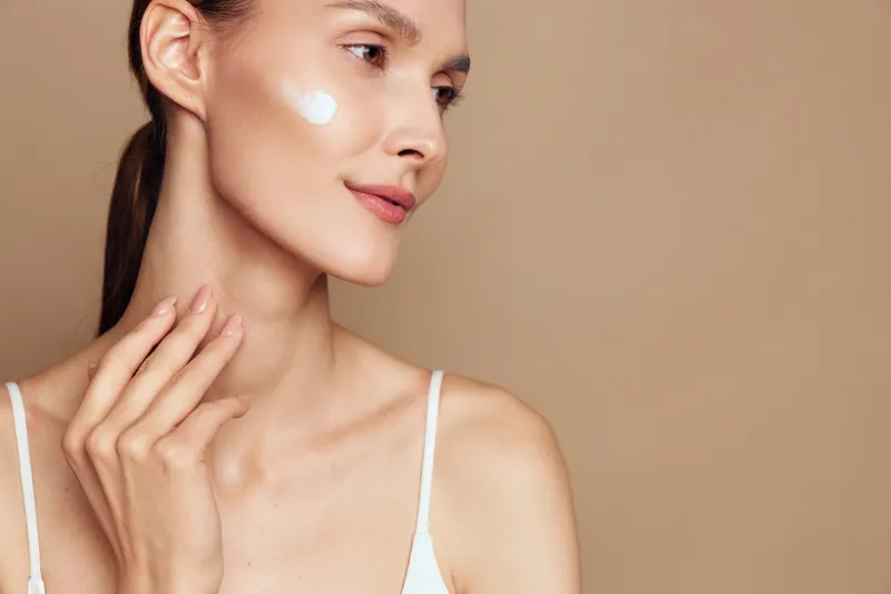 Young woman with clear skin applying white face cream on cheek. Close-up portrait on beige background. Concept of skincare, beauty, wellness, and self-care with natural lighting.