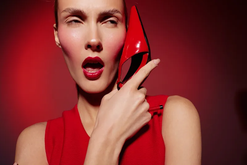 Close-up of a woman in red dress holding a red high heel shoe near her face with expressive makeup and glossy lips. Concept of fashion, beauty, and bold style.