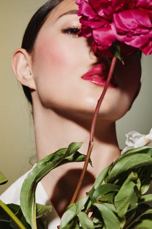 Close-up of a woman with vibrant makeup holding pink flower partially covering her face. Concept of beauty creative fashion, color contrast, elegance and style.