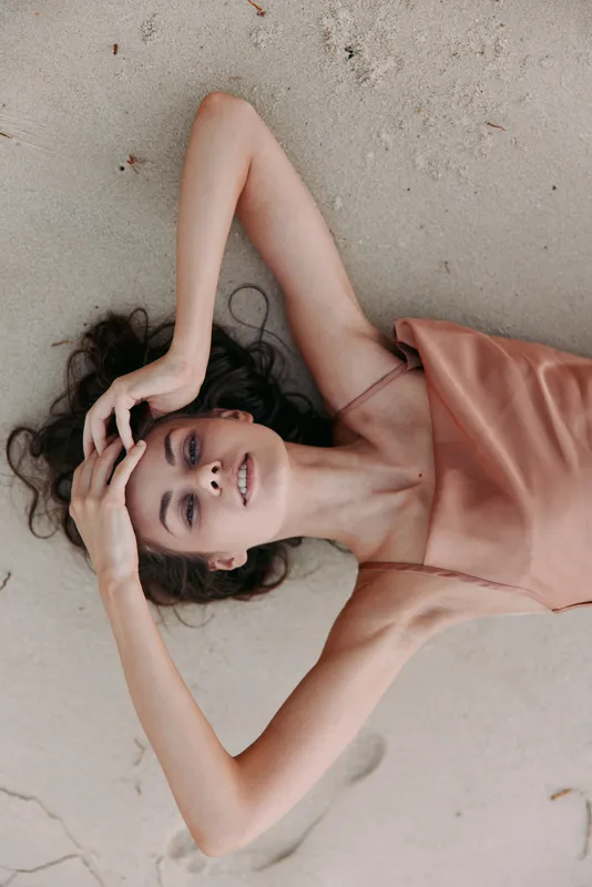 Portrait of a young woman lying on sandy beach, wearing a peach satin dress, relaxed pose, smiling softly, natural light, candid mood, graceful lines and tranquil vibe.