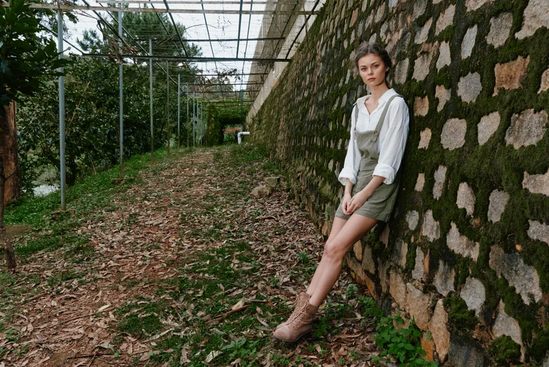 Young woman in green overalls against a stone wall in a lush forest environment, exuding a calm and serene vibe, embodying nature and seasonal beauty