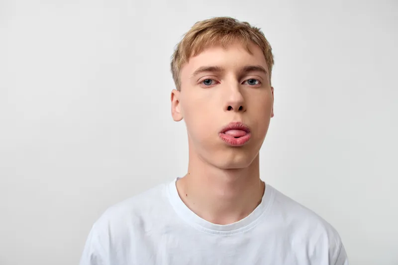 Young man with playful face sticking tongue out against a plain white background, casual wear and relaxed mood, close up facial expression in studio