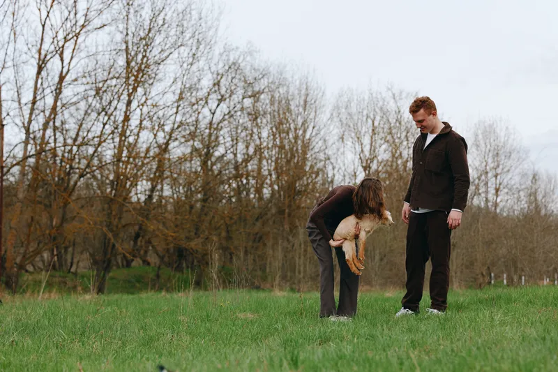 Young couple spends time outdoors in early spring while woman holds a small goat in her arms and man watches on a grassy field with bare trees behind them.