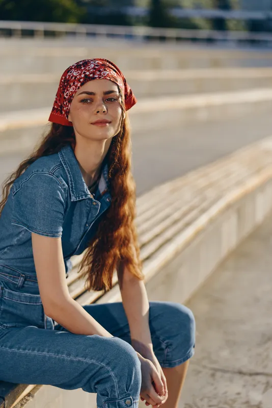 Young woman with long hair wearing a red bandana and denim outfit sitting outdoors in warm sunlight with relaxed expression and casual style.