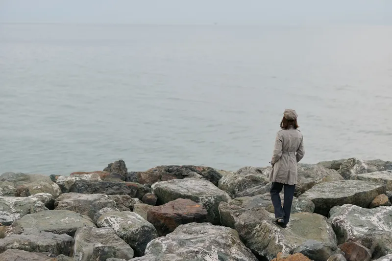 A woman in a grey coat stands alone on rocky shore facing calm sea. Overcast sky and muted colors create a contemplative atmosphere of solitude and reflection by the water.
