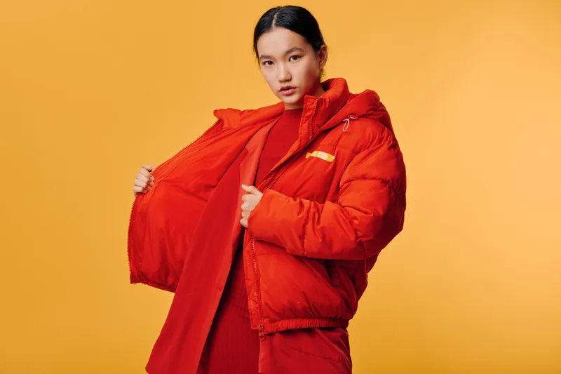 Young woman wearing bright red winter coat and matching sweater posing against solid orange background for fashion and seasonal style portrait, casual warm clothing and confident look.