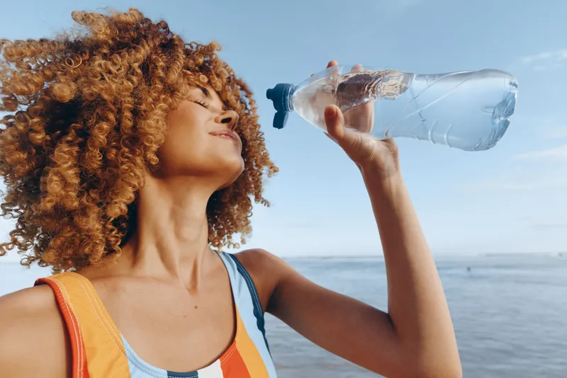 Woman with curly hair drinking water from a plastic bottle by the ocean on a summer day, closeup profile outdoors, sunlight, thirst relief, hydration and wellbeing, fresh air breeze today
