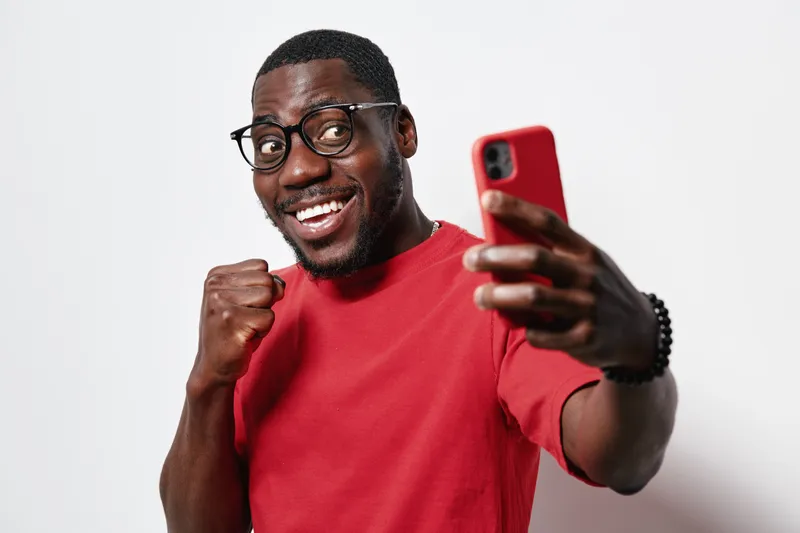 African American man with dark skin and glasses wearing red shirt taking selfie with smartphone smiling and showing fist gesture on white background.