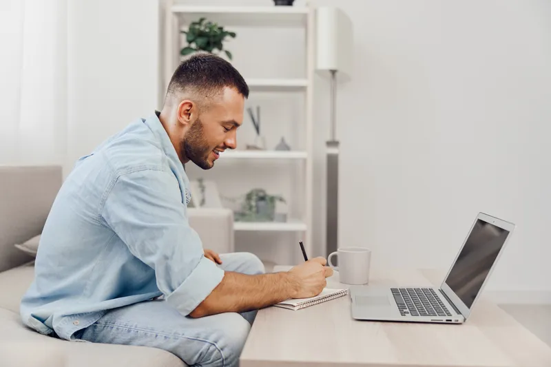 Focused man working on a laptop while taking notes in a modern, minimalistic home office. Bright and inviting atmosphere that inspires productivity.