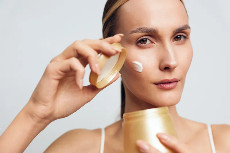 Young woman applying face cream with soft texture on cheek, natural skin care routine, neutral background, focused expression, beauty treatment at home