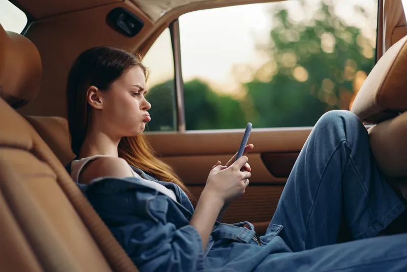 Young woman using smartphone in car, expressing frustration or boredom with denim outfit and natural background, perfect for lifestyle and travel themes.