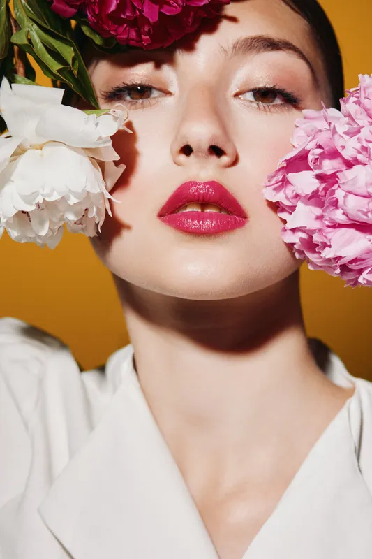 Young woman with glossy red lips and flawless skin poses with pink and white flowers near face against yellow background, creating artistic beauty and fashion concept.