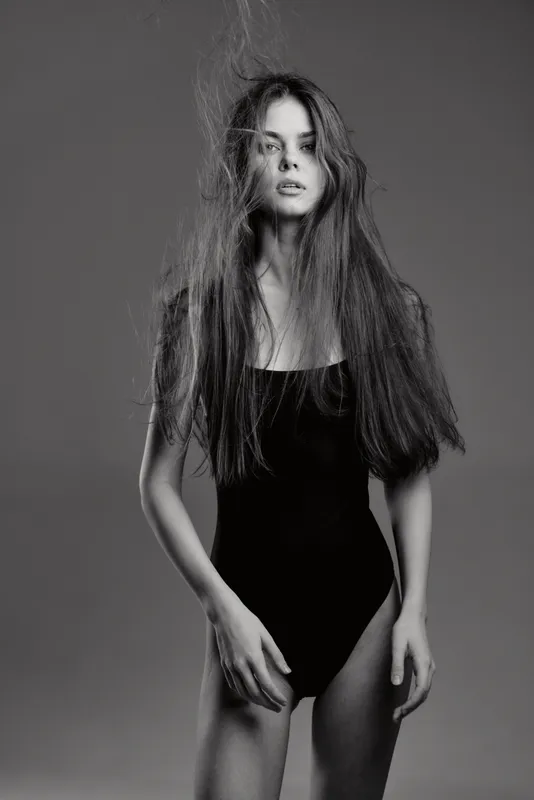 Black and white portrait of a young woman with long hair wearing a black bodysuit, posing confidently with neutral expression.