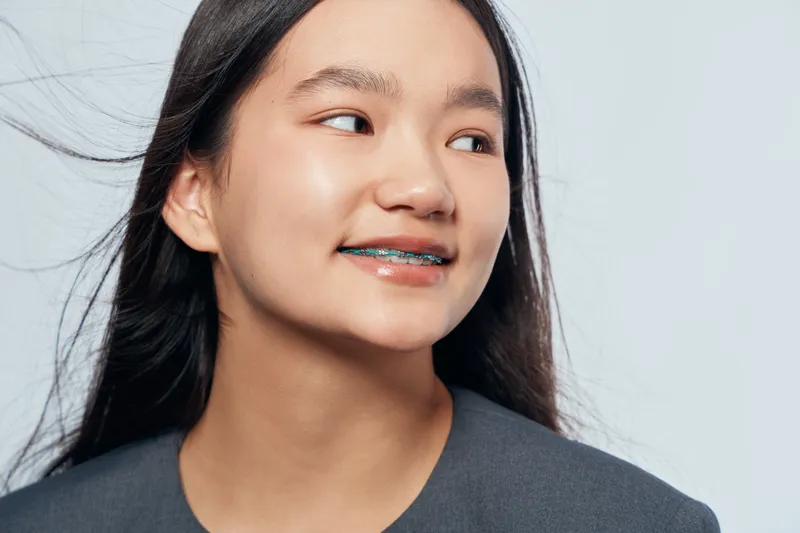 Teenage girl with braces smiling and looking aside against plain background. Portrait of young female with dental appliance and long dark hair in casual clothes indoors.