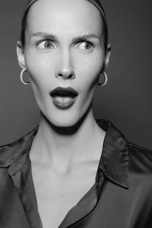 Surprised white woman with pale skin and slicked hair wears hoop earrings and satin shirt. Studio portrait in black and white with dramatic expression.