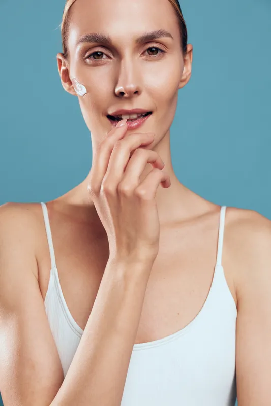 A young woman in a white tank top applies cream to her face with a gentle smile, showcasing skincare routine and natural beauty on a soft blue background.