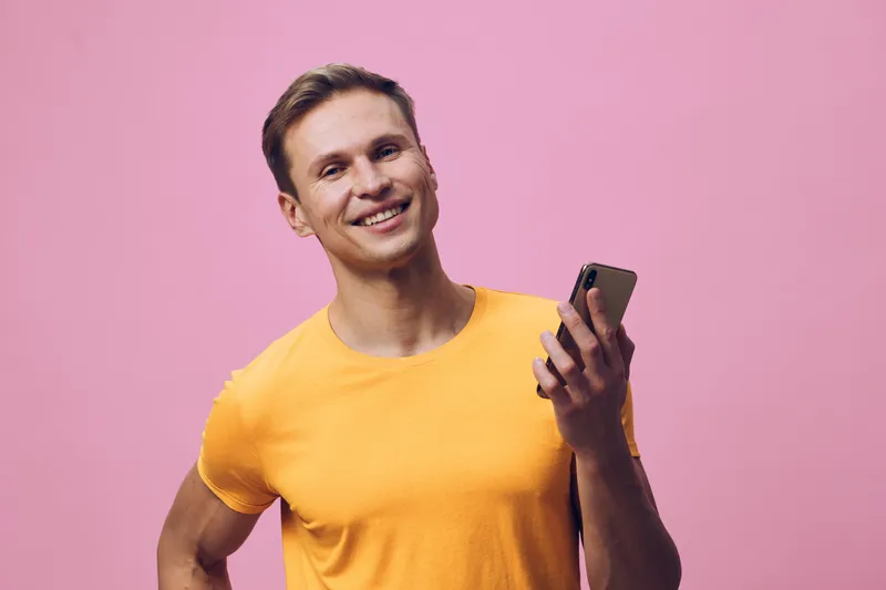 smiling young caucasian man wearing yellow t-shirt holding smartphone look camera isolated on pink background digital nomad remote work concept