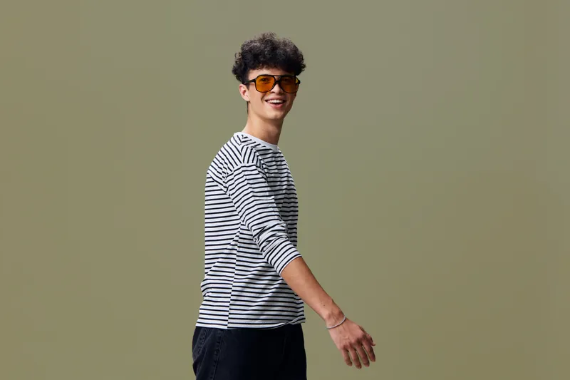 Smiling man wearing striped shirt and sunglasses stands against olive background expressing casual style and positive mood with curly hair and relaxed posture.