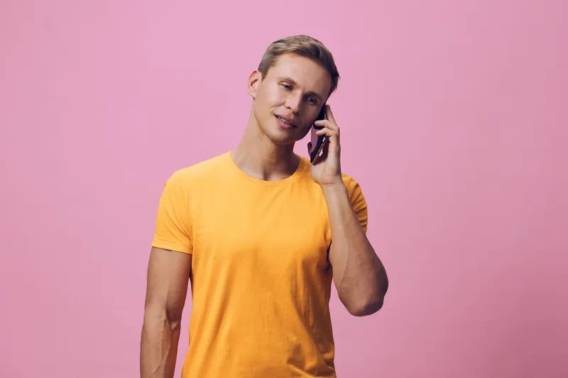 Smiling man talking on smartphone casual young adult wearing yellow t-shirt isolated on pink background portrait. Lifestyle concept.