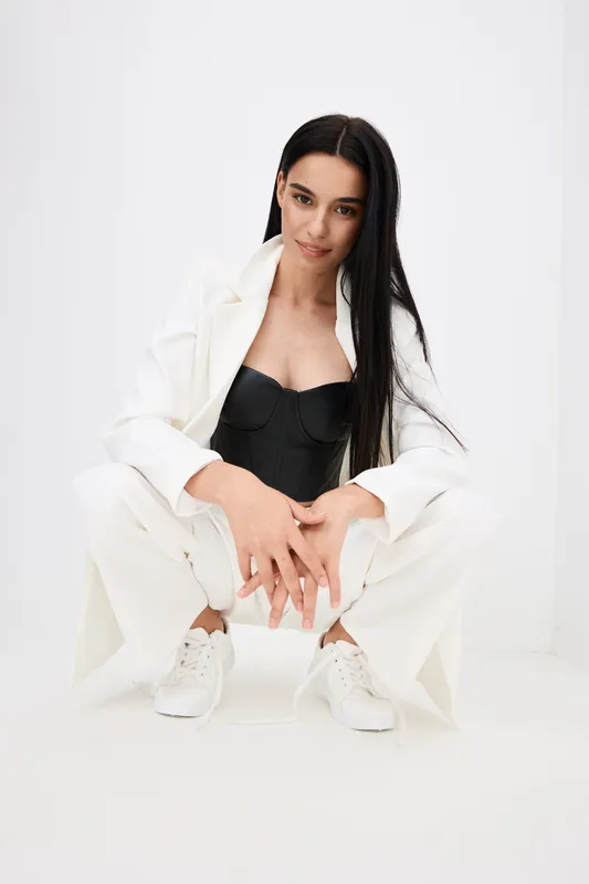 young woman in white suit squatting, wearing black corset and white sneakers, posing on white background with relaxed confident expression