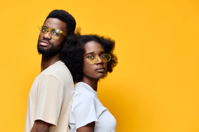 Stylish young couple with curly hair in trendy glasses against vibrant yellow background, showcasing contemporary fashion and confident emotions