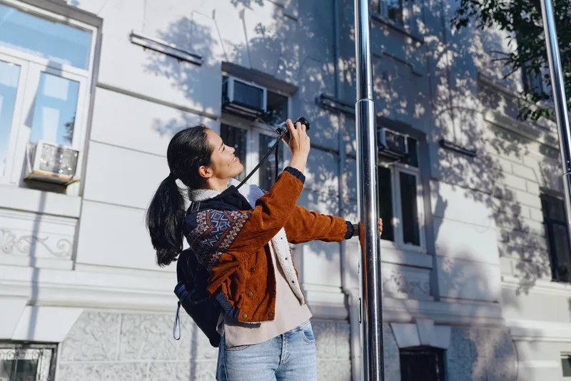 Photographer outdoors capturing a shot on a sunny urban street, handheld camera and phone raised toward a metallic pole, backpack on back, casual jacket, focused expression