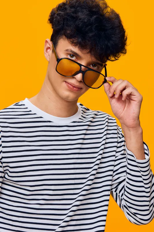 young man wearing striped shirt and amber sunglasses looking playfully against vibrant orange background with curly hair and casual style