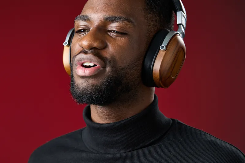 Young Black man enjoying music with wooden headphones, wearing a black turtleneck, against a vibrant red background, expressing joy and relaxation