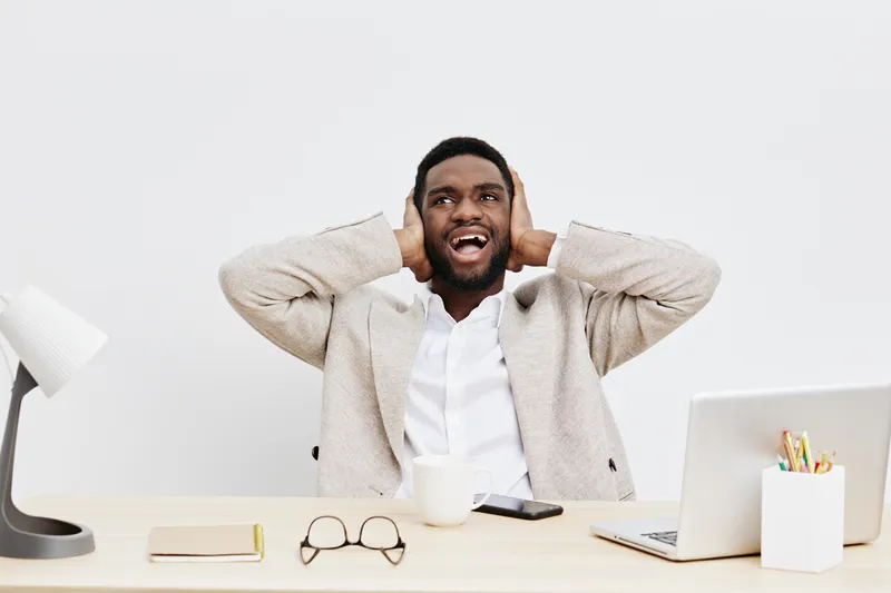 Happy young african american man sitting at desk with a laptop, wearing a beige blazer and white shirt, covering ears with hands, surprised and amused, modern workspace, bright background, casual offi