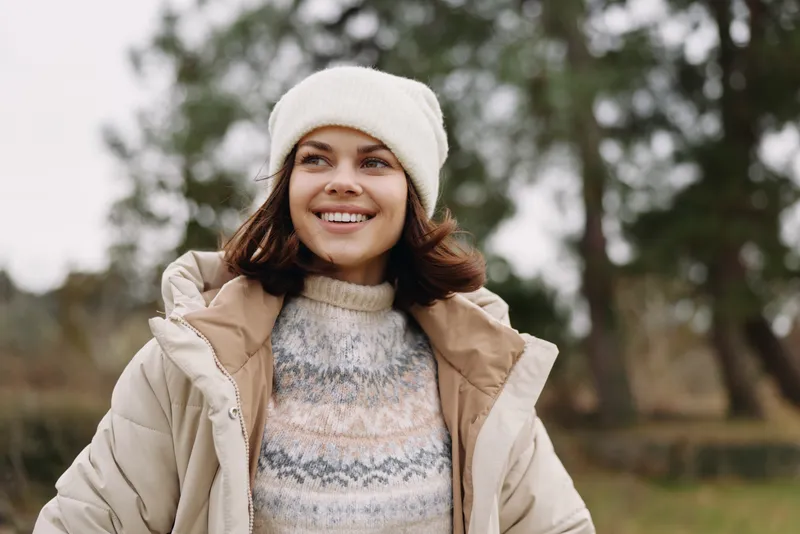 Woman outdoors wearing a warm coat and beanie, smiling in a park setting with trees. Casual winter fashion shot highlighting cozy outerwear and approachable style for lifestyle or ecommerce use.