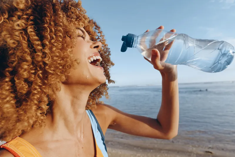Woman, curly, hair, drinking, water, sea, summer, hydration, lifestyle, outdoors, smiling closeup of female, plastic bottle in hand, clear refreshing drink, ocean horizon, warm sunlight, wellbeing