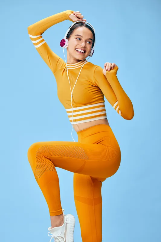 Active woman in bright orange fitness outfit dancing with headphones, showcasing high energy movement, athletic flow and joyful workout vibe in a clean studio backdrop.
