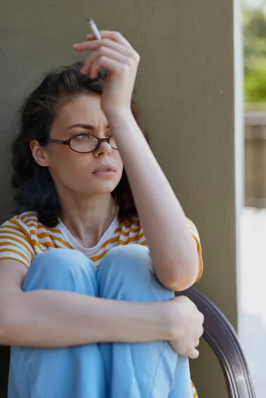 A thoughtful woman in a striped shirt sits on a porch, holding a cigarette and looking down with a pensive expression, conveying quiet contemplation and solitude.