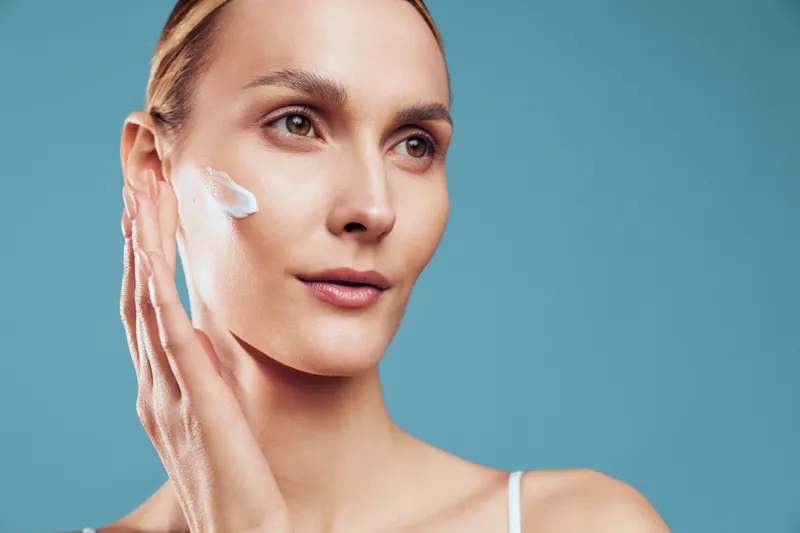 A young woman applies moisturizing cream to her cheek with her hand against a plain blue background. The image represents skincare, beauty routine, and healthy skin care concepts.