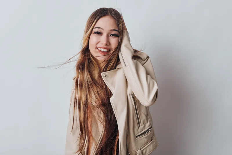Young woman with long hair wearing beige jacket posing and smiling on white studio background. She looks confident, happy and relaxed with natural makeup and casual style.