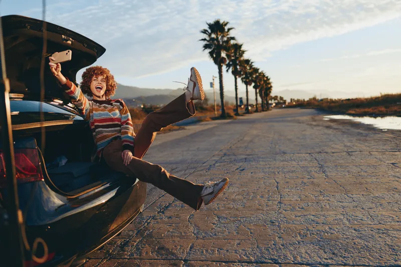 Happy young woman by open car door on long road at sunset, laughing with legs out, carefree lifestyle travel adventure, palm trees and highway outdoors background.