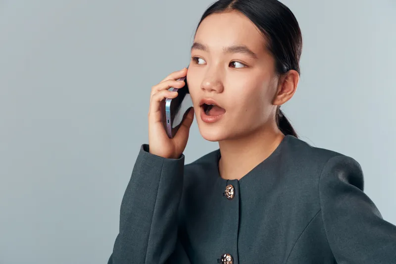 Young woman in business attire talking on smartphone with surprised expression on face isolated on light background. Female professional communicating with mobile device for work or personal use.