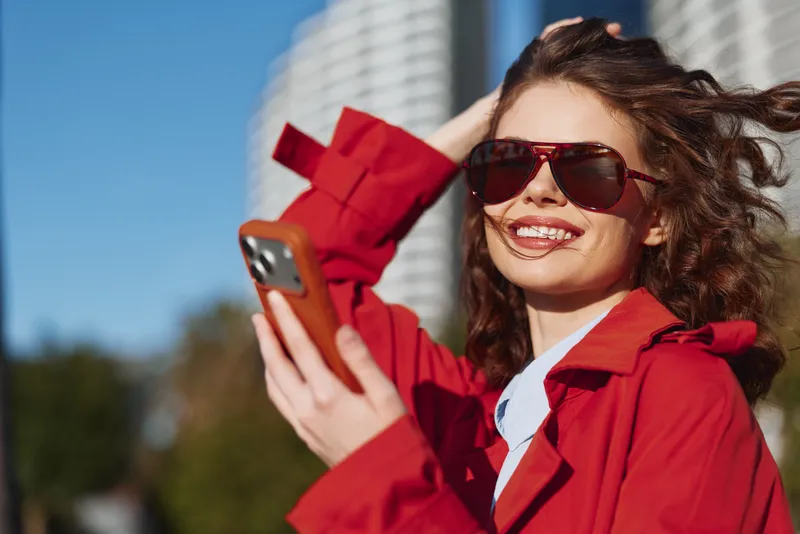 Young woman in a red jacket takes a selfie outdoors with sunglasses on, a sunny city backdrop, and a casual, breezy vibe that captures a playful, candid moment.