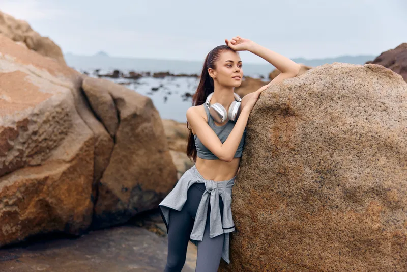 Athletic young woman in sportswear poses by rocky beach, showcasing fitness lifestyle and healthy living, wearing headphones and expressing confidence