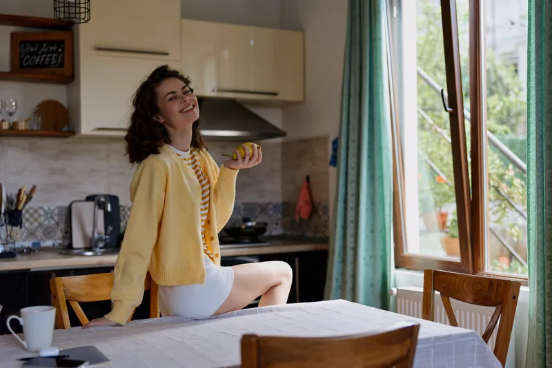 Cheerful woman indoors in a bright kitchen, casual outfit with a yellow cardigan, enjoying a sunny breakfast moment, relaxed posture, natural colors, cozy interior. soft breeze glow
