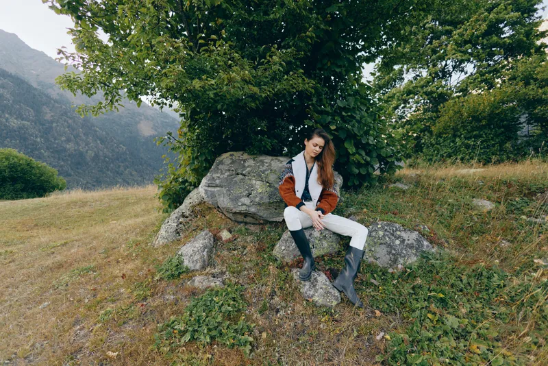 Woman sitting on rock under tree in mountain field outdoors wearing fashionable coat and boots, relaxed portrait and countryside nature scene with scenic mountains and calm mood